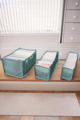 Ahşap zeminde üç farklı boyutlu mavi kumaş cepli düzenleyici, içlerinde katlanmış giysilerle