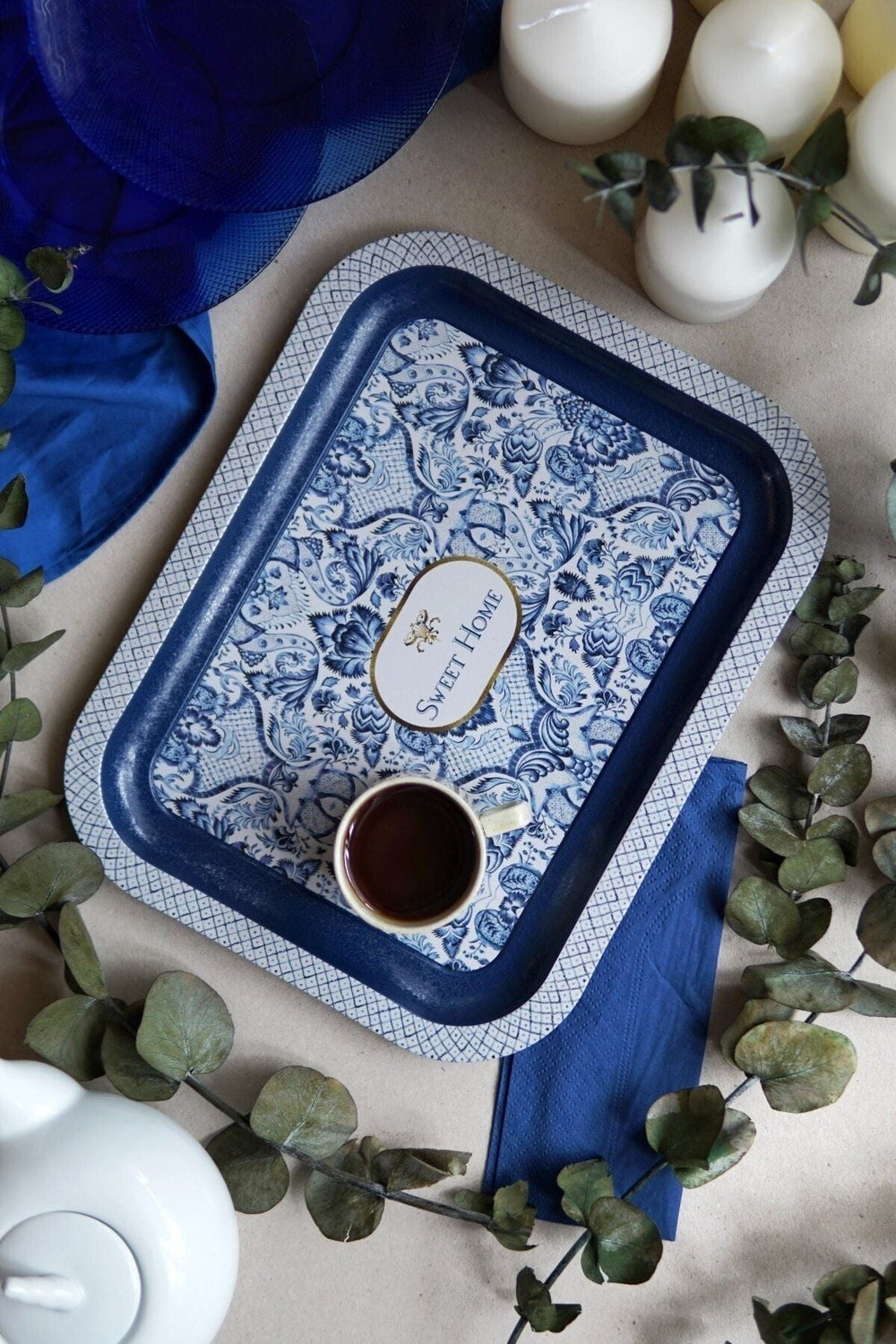 Mavi desenli Sweet Home tepsi üzerinde kahve fincanı, beyaz mumlar ve yeşil yapraklar