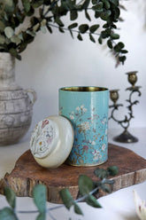 Mavi çiçek desenli dekoratif teneke kutu, ağaç dilimi üzerinde, arka planda antika vazo ve şamdan