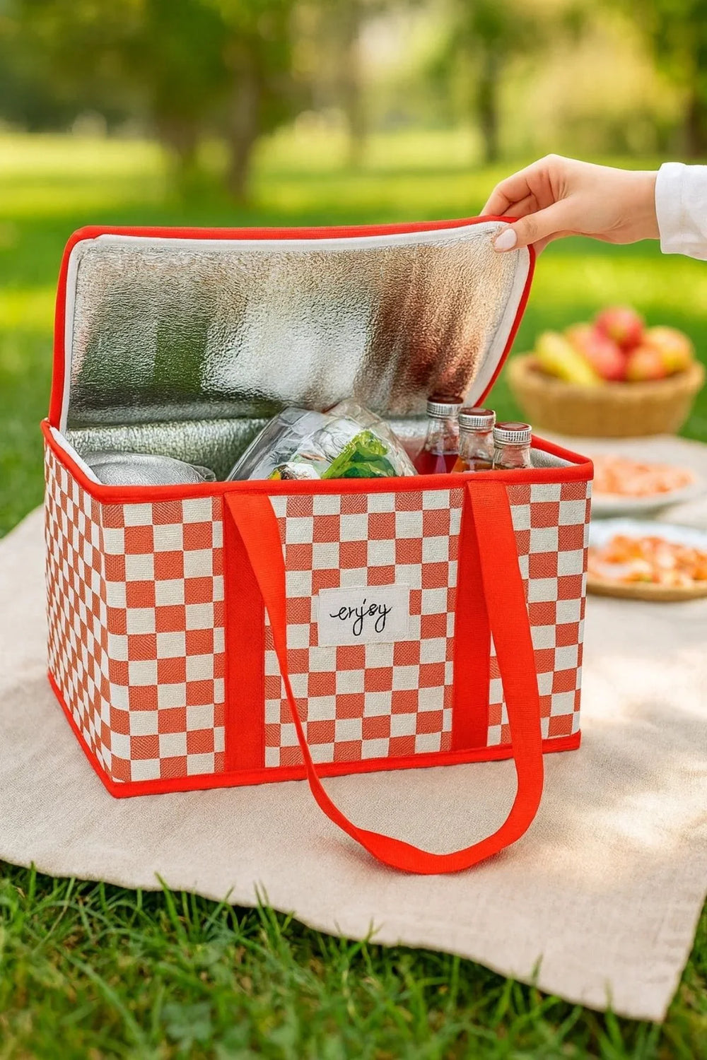 Kırmızı beyaz kare desenli yalıtımlı piknik çantası, açık kapağı ve içindeki yiyecek ile yeşil park ortamında