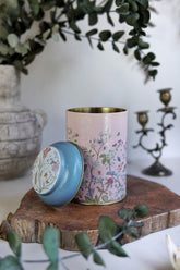 Ahşap yüzeyde üzerinde mavi kapaklı, çiçek desenli metal kutu, arka planda vazo ve şamdanlar