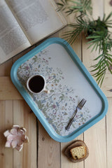 Mavi kenarlıklı çiçek desenli tepsi üzerinde kahve fincanı ve çatal, yanında kek dilimi ve açık kitap