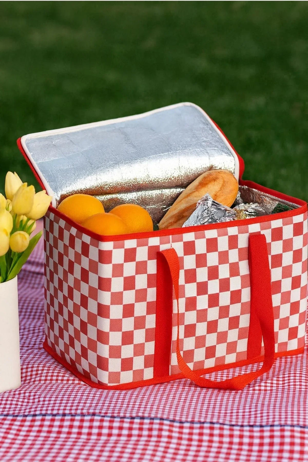 Kırmızı beyaz kare desenli soğutucu çanta içinde ekmek, mandalina ve folyo paketler, kırmızı ekose örtü üzerinde