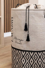 Bej ve siyah desenli, püsküllü kumaş çamaşır sepeti, iç mekan ahşap zemin