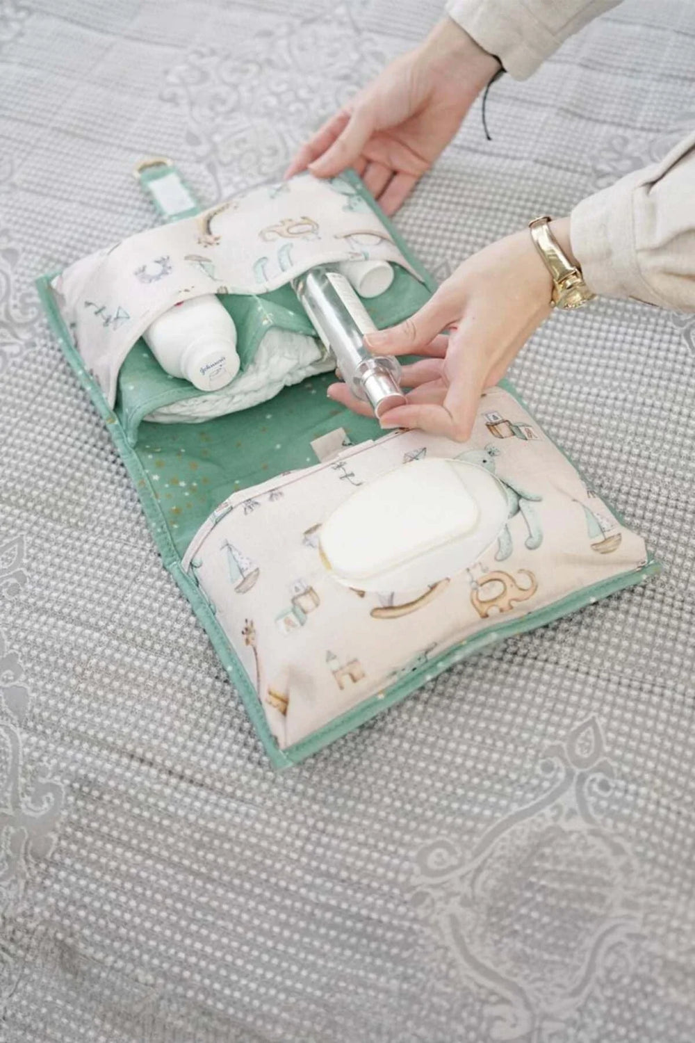 Bebek bakım çantası içinde krem ve su şişesi bulunan, gri örtü üzerinde açılmış bebek bakım seti