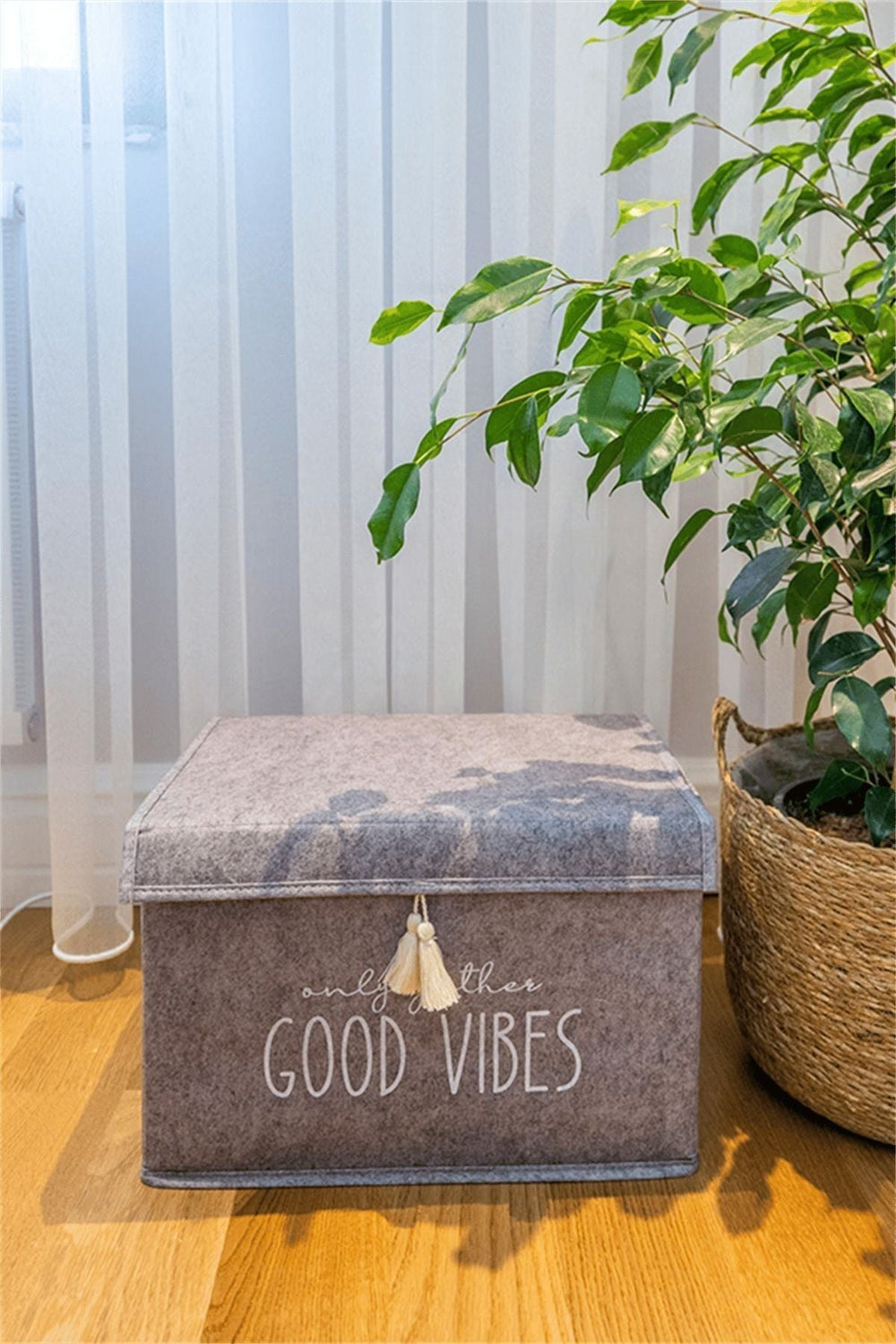 Katlanabilir gri kutu üzerinde 'Only good vibes' yazısı, yanındaki büyük saksı bitkisi ve ahşap zemin