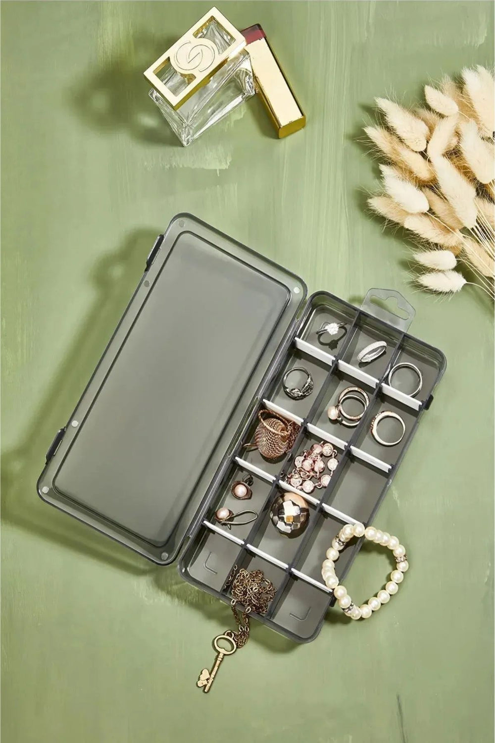 Yeşil zeminde açık plastik takı kutusu, içindeki yüzükler, küpeler ve inci bileklik ile parfüm şişesi ve dekoratif kuru bitkiler