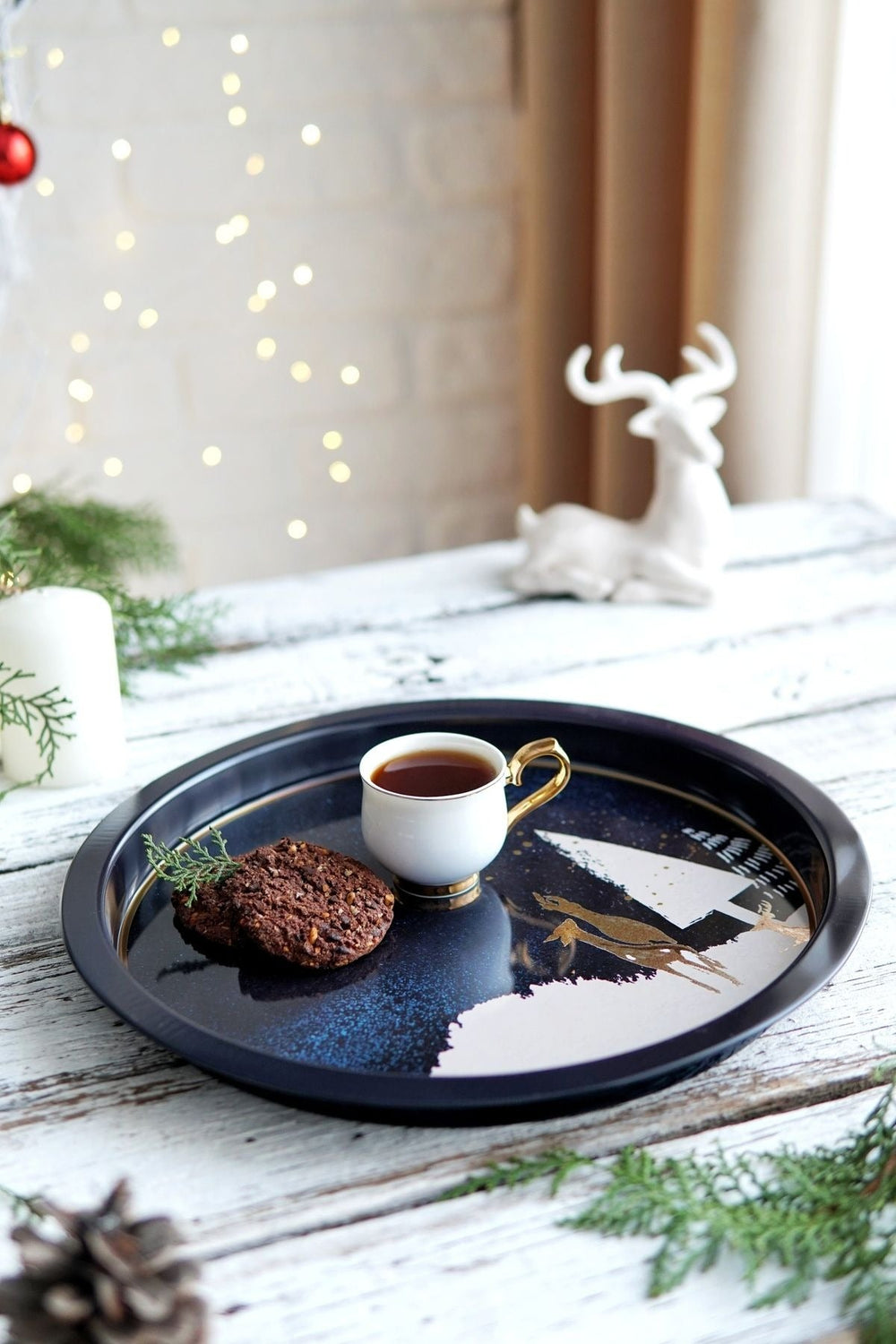 Kış temalı tepside kahve fincanı ve çam dallı iki kurabiye, arka planda beyaz geyik figürü