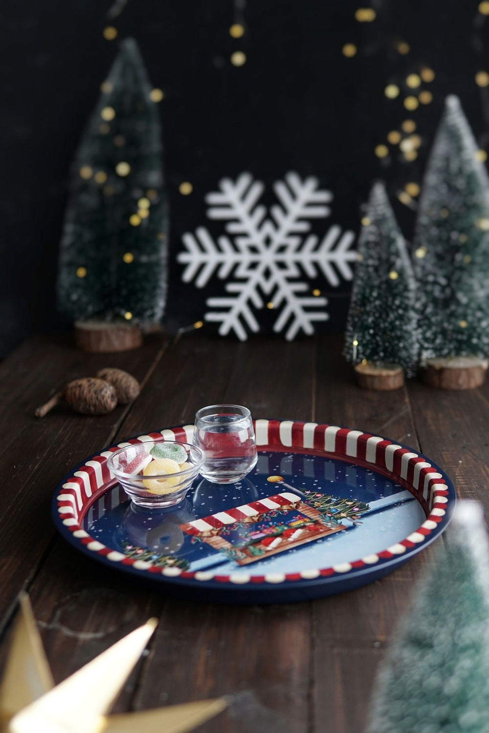 Kahverengi ahşap masada, kar desenli tepsi içinde küçük pipetli su bardağı ve renkli jelatinli şekerler