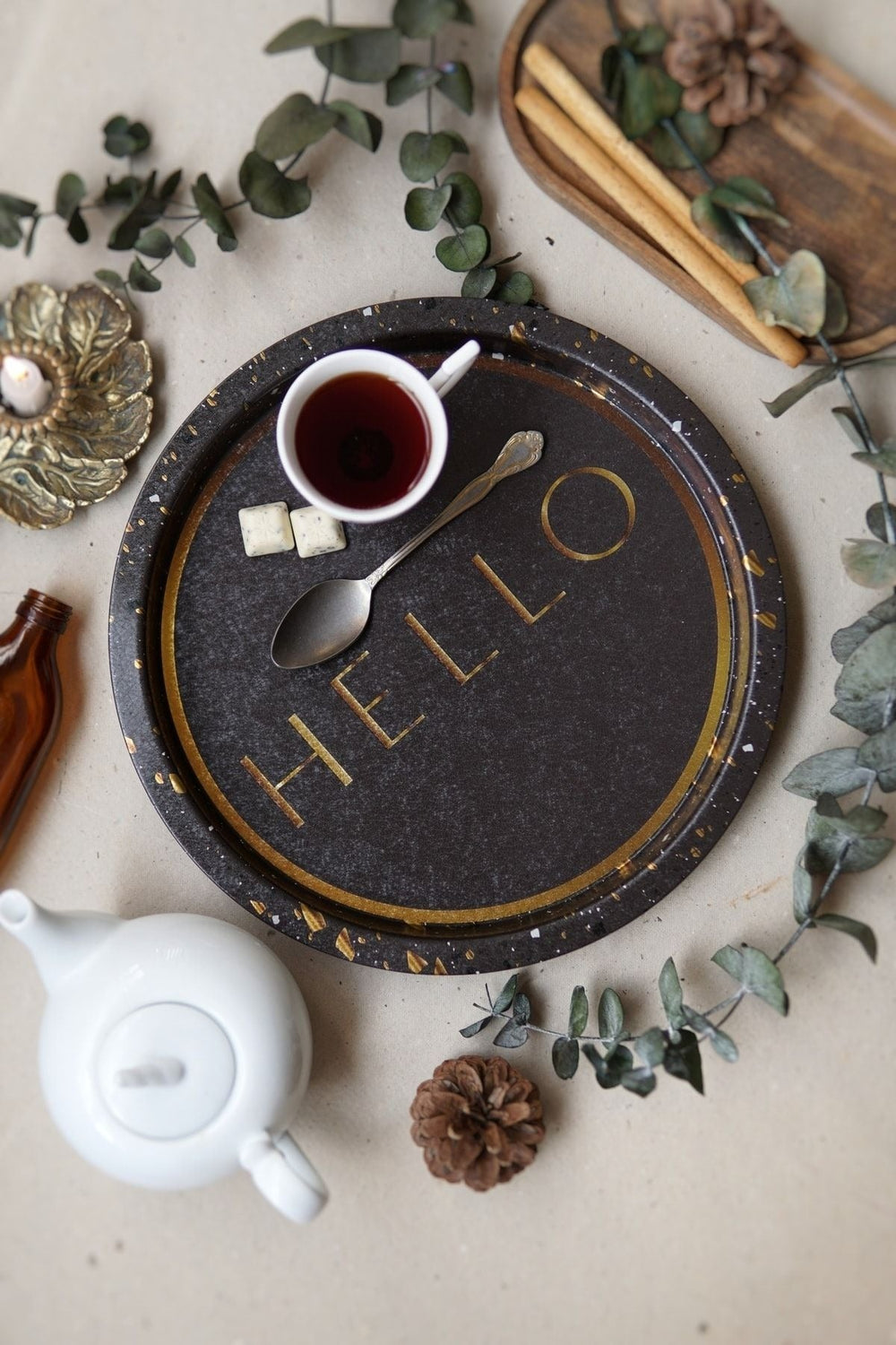 Kahverengi tepsi üzerinde beyaz kupa içinde siyah çay, iki beyaz şeker, gümüş kaşık, beyaz çaydanlık, dekoratif bitkiler ve tarçın çubukları