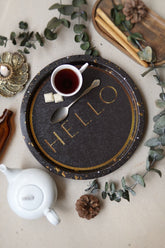 Kahverengi tepsi üzerinde beyaz kupa içinde siyah çay, iki beyaz şeker, gümüş kaşık, beyaz çaydanlık, dekoratif bitkiler ve tarçın çubukları