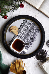 Beyaz zeminde kitap, siyah kenarlı tepside çay ve bisküvi, kurutulmuş çay yaprakları ve yeşil yapraklı kırmızı meyveler