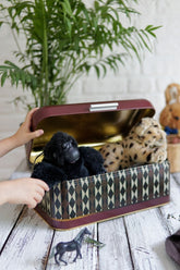 Elde tutulan siyah peluş goril ve leopar desenli kutuda oyuncaklar, ahşap masa ve yeşil bitki arka planda