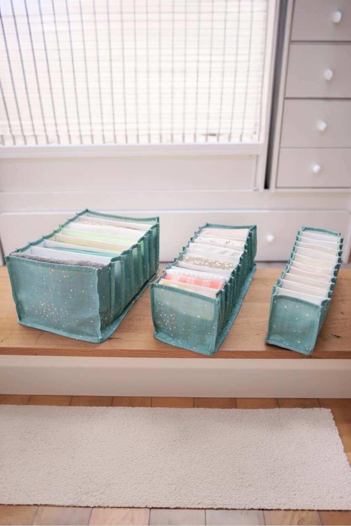 Ahşap zeminde üç farklı boyutlu mavi kumaş cepli düzenleyici, içlerinde katlanmış giysilerle