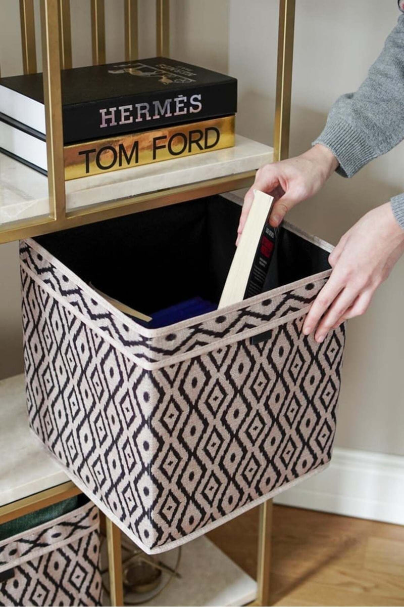 Geometrik desenli saklama kutusuna kitap yerleştiren kişinin elleri, raf üzerinde Tom Ford ve Hermès kitapları