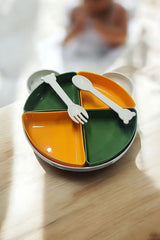 Yeşil ve turuncu bölmeli çocuk tabağı, üzerinde beyaz kemik şeklinde kaşık ve çatal