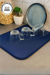 Mavi mutfak bezi üzerinde ters çevrilmiş cam bardaklar ve tabaklar, arka planda beyaz duvar ve bitki