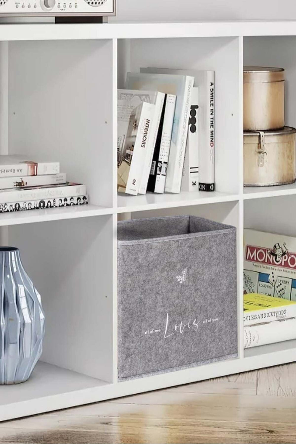 Beyaz kitaplıkta gri saklama kutusu, kitaplar, dekoratif mavi vazo ve metal kutular