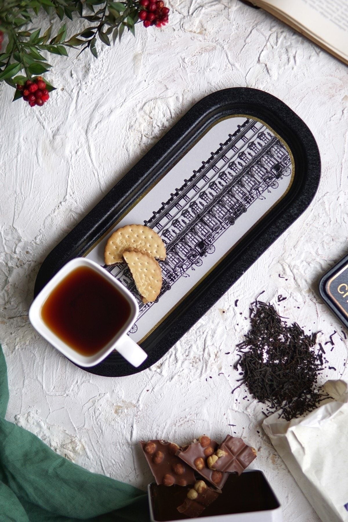 Beyaz zeminde siyah tepside çay, bisküvi, porselen fincan ve sütlü fındıklı çikolata parçaları