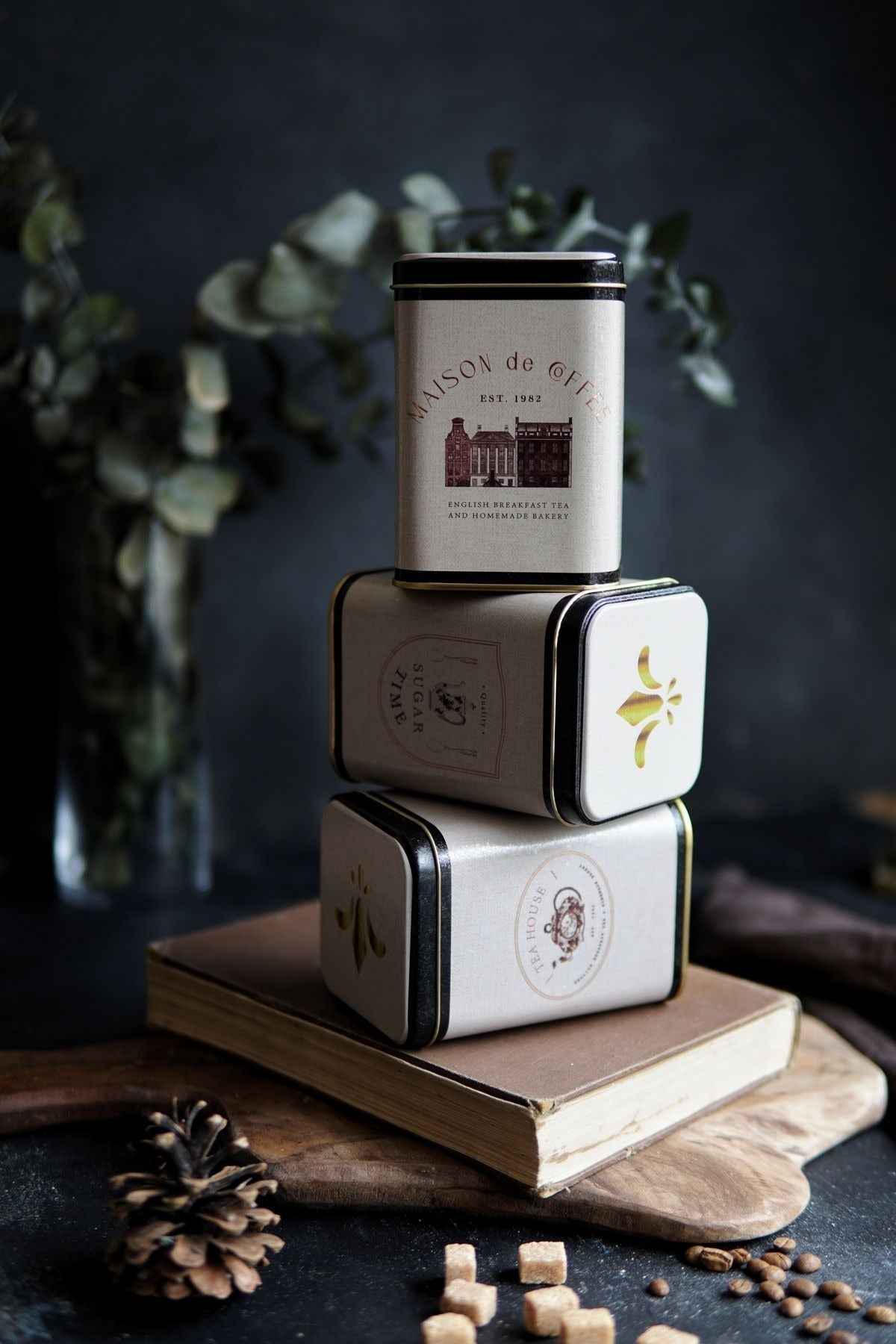 Kitap üzerinde üst üste dizilmiş Maison de Coffee, Sugar Time ve Tea House metal kutular, arka planda kuru yaprak dekor