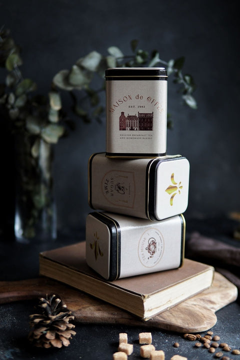 Kitap üzerinde üst üste dizilmiş Maison de Coffee, Sugar Time ve Tea House metal kutular, arka planda kuru yaprak dekor