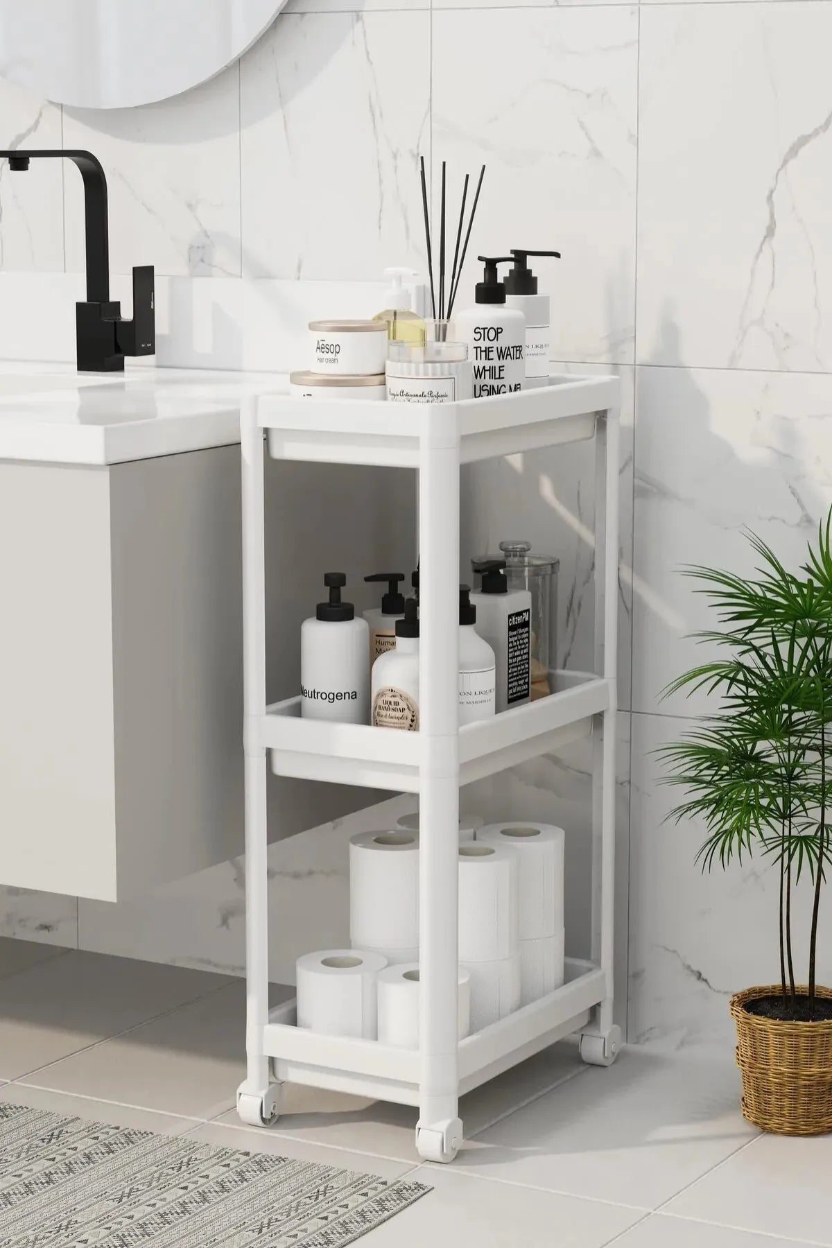 Beyaz çok katlı tekerlekli banyo rafında tuvalet kağıdı ve bakım ürünleri, mermer duvar, siyah musluk ve saksıda bitki