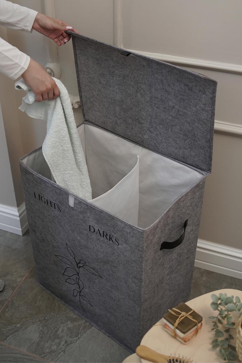 Gri iki bölmeli kumaş çamaşır sepeti, biri açık renkler diğeri koyu renkler için etiketli, havlu yerleştiriliyor
